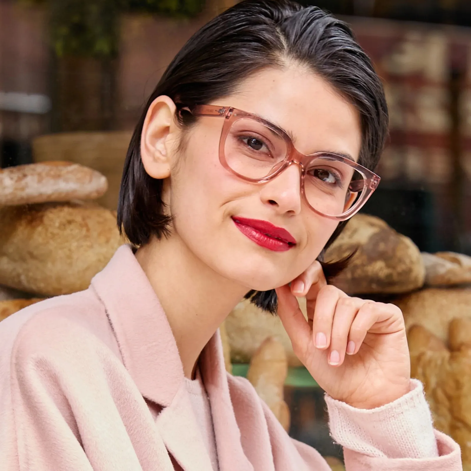 A woman with a sleek dark bob and red lipstick in a blush coat, hand at her chin in front of a bakery window, wearing LOOK OPTIC Evita readers in Shiny Pink