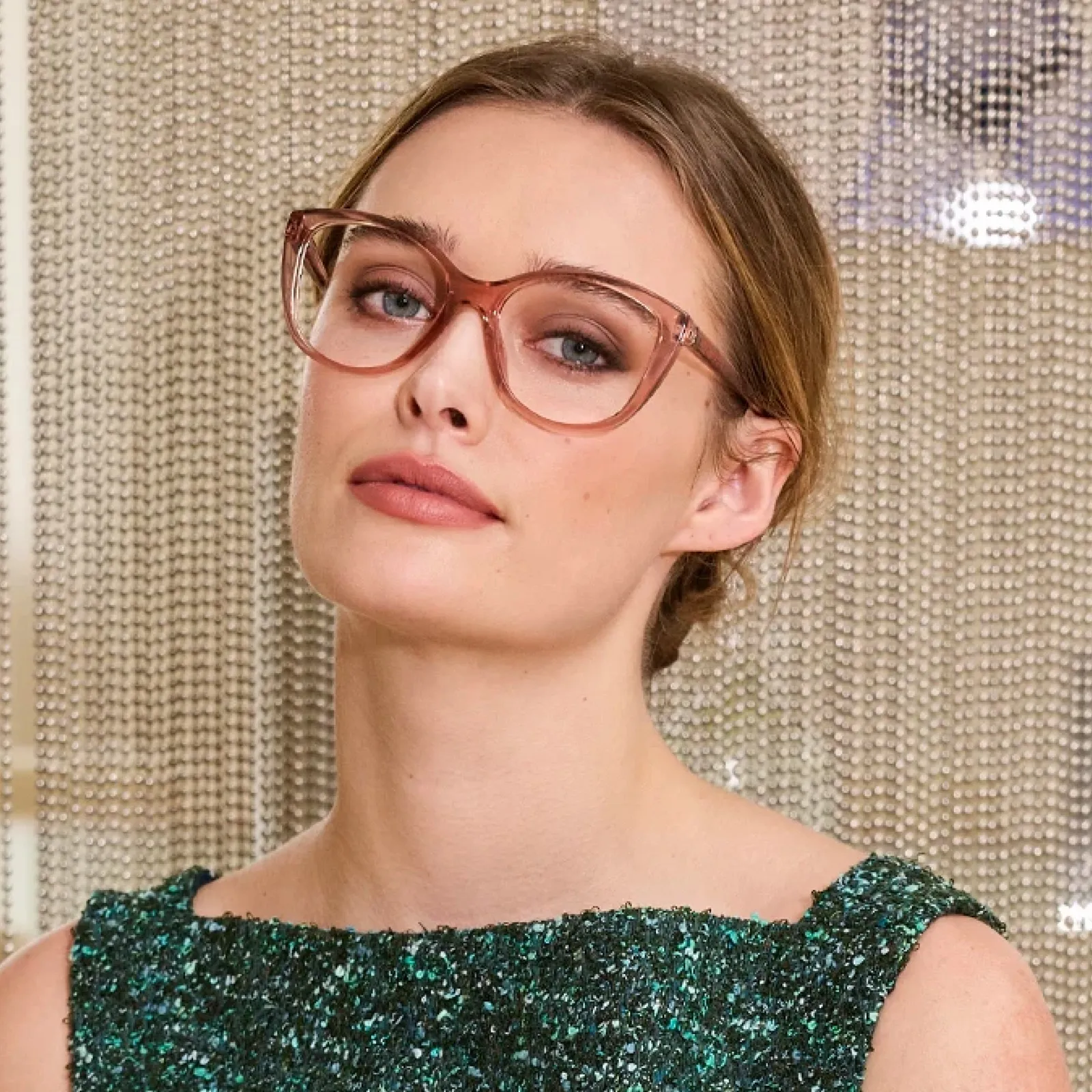 A woman with her hair pulled back in a green sparkle-tweed top in front of a beaded curtain, wearing LOOK OPTIC Keira readers in Shiny Pink